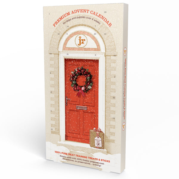 JR Pure Dog Advent Calendar | Christmas Dog Treats Venison & Turkey. A boxed advent calendar package standing upright on a white studio background. The front shows an illustrated red front door with a holiday wreath, arched doorway details, and red text reading 'PREMIUM ADVENT CALENDAR' and 'for dogs and puppies over 4 weeks. 