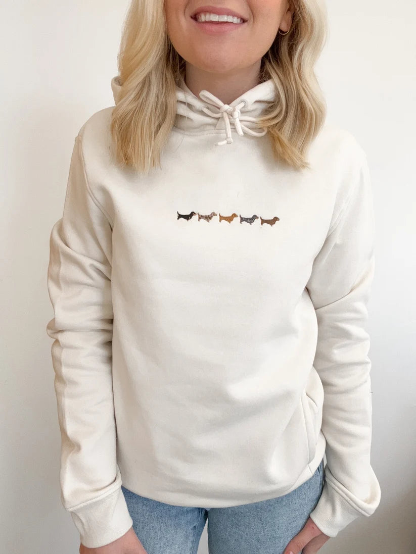 A woman wearing a cream-colored hoodie with a drawstring hood and a small embroidered row of dachshund dogs across the chest, paired with light blue jeans.
