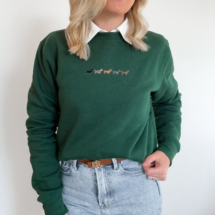 A woman wearing a dark green crewneck sweatshirt with a small embroidered line of colorful dogs on the chest, styled with a white collared shirt and light-wash jeans.