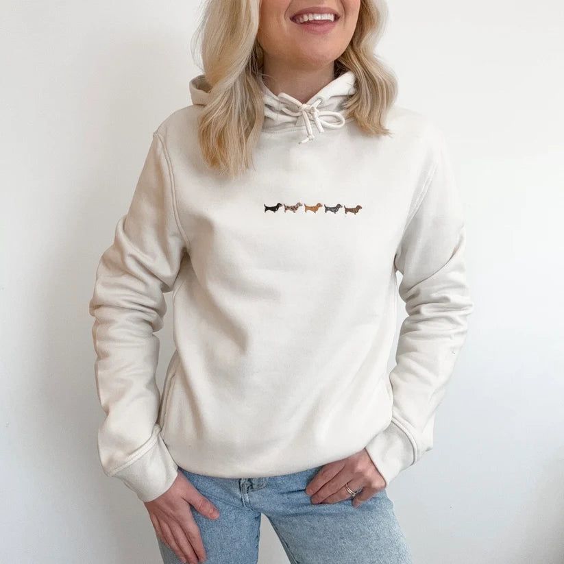 A woman wearing a cream-colored pullover hoodie with a small embroidered row of dachshunds across the chest, paired with light blue jeans, set against a plain white wall.