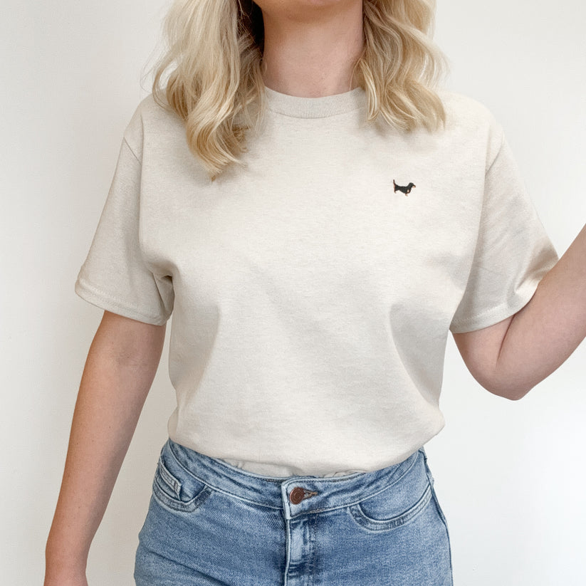 A woman wearing a cream-colored short-sleeve crewneck t-shirt with a small black dachshund logo on the left chest, paired with blue jeans.