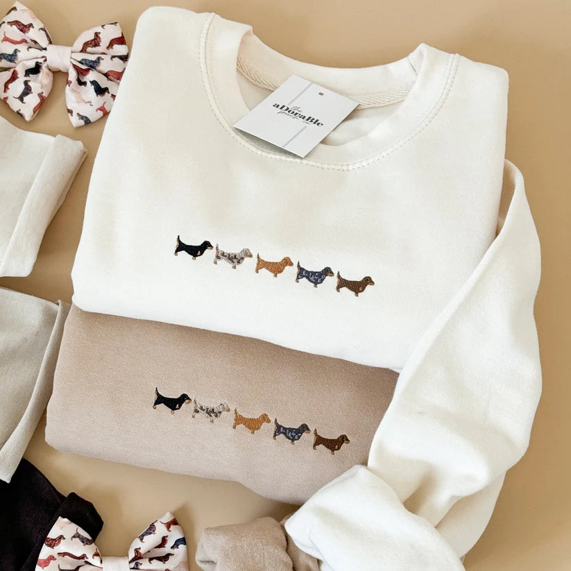 A folded cream-colored long-sleeve sweatshirt with a row of embroidered dachshund dogs across the chest, displayed on a beige background alongside other folded garments and patterned bows.