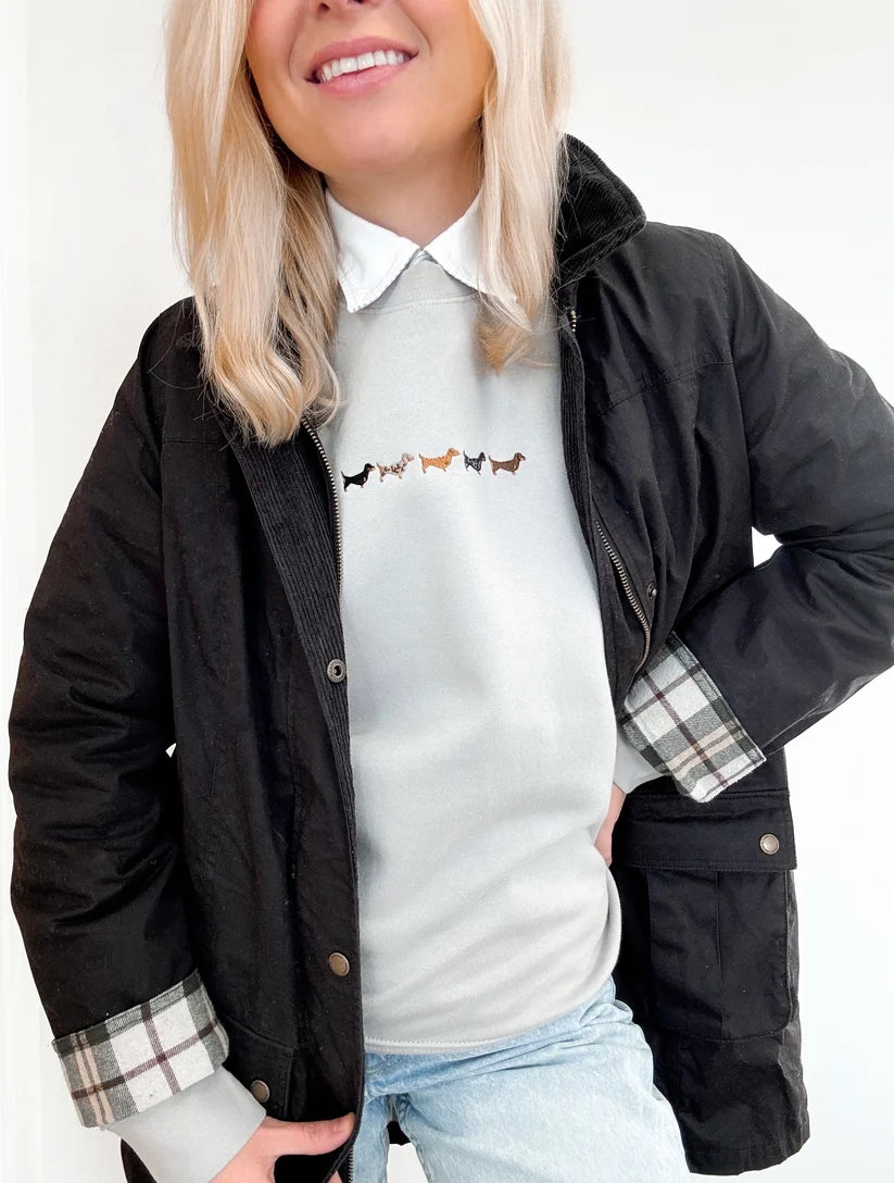 A woman wearing a black waxed cotton jacket over a white collared sweater with a small dog line graphic, paired with light blue jeans. The jacket features a plaid lining on the cuffs and a zippered front.