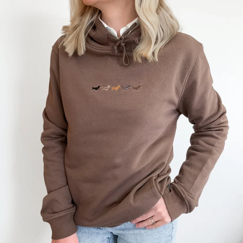 A woman wearing a brown cowl neck sweatshirt with a small embroidered line of dachshunds across the chest, paired with light blue jeans against a plain white background.