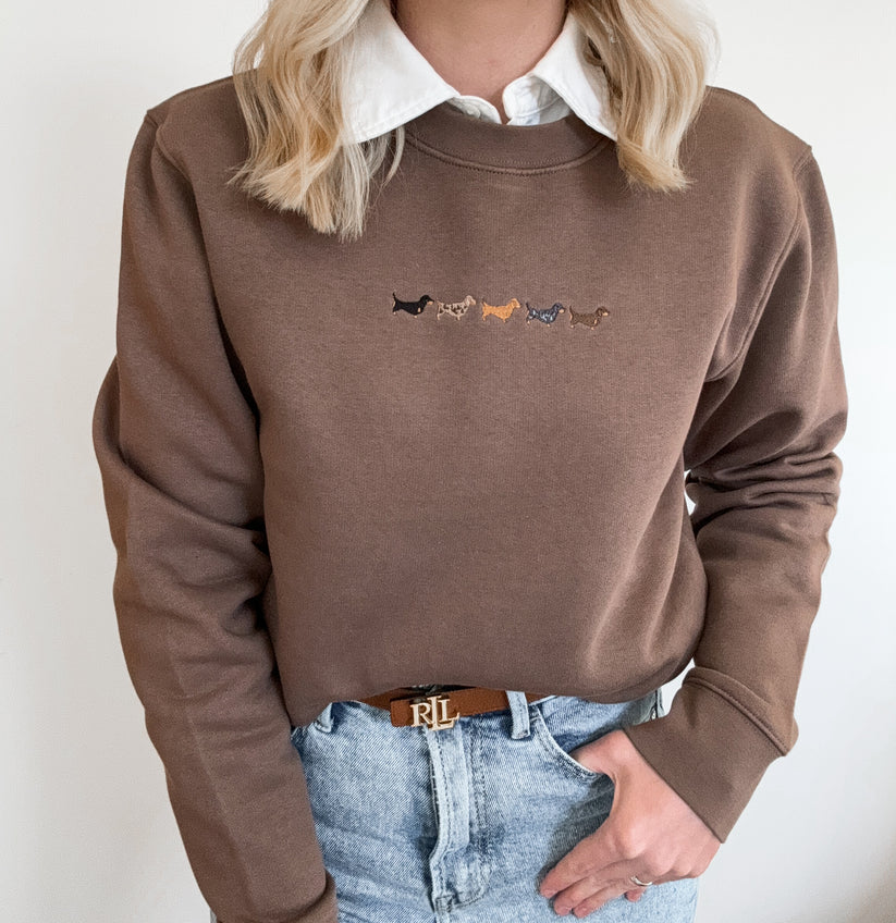 A person wearing a brown crewneck sweatshirt with a white collared shirt underneath and light blue jeans, featuring a small embroidered line of dachshunds on the chest.