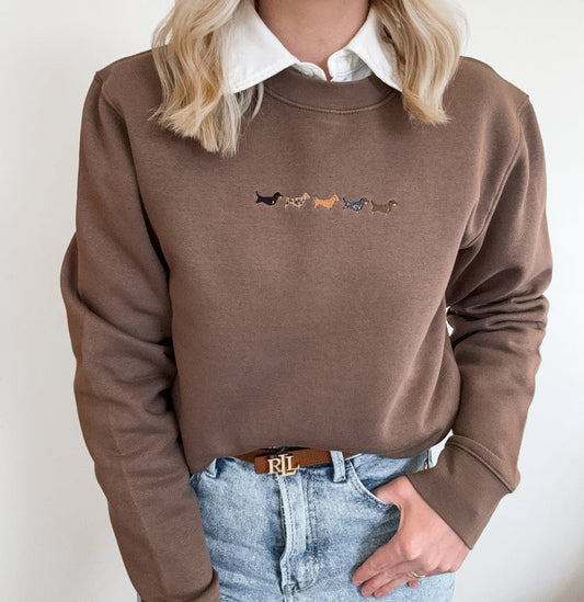 A person wearing a brown crewneck sweatshirt with a white collared shirt underneath and light blue jeans, featuring a small embroidered line of dachshunds on the chest.