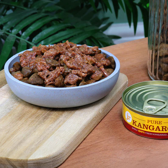 A bowl of wet pet food sits on a wooden cutting board next to an open can of Pure Kangaroo wet dog food on a wooden table, with green leaves in the background.