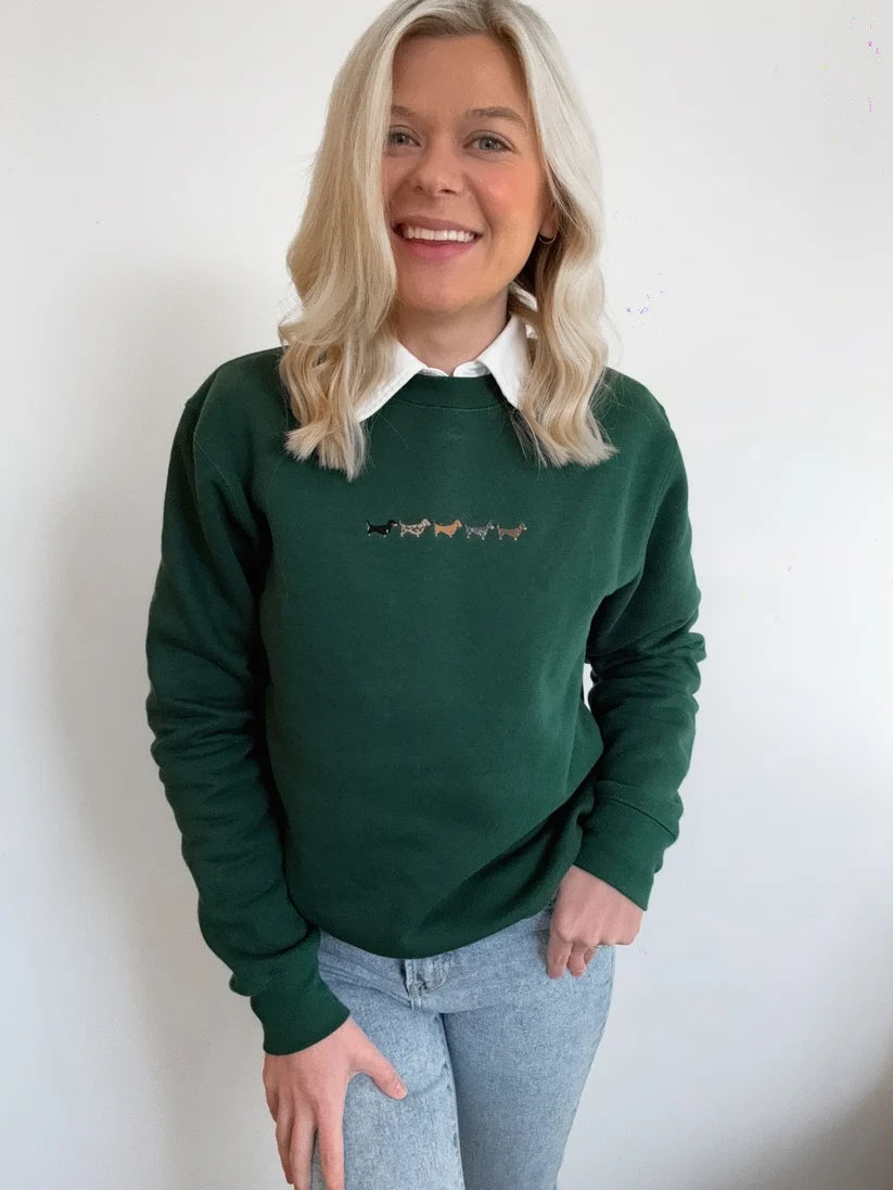 A smiling blonde woman wearing a dark green crewneck sweatshirt with a small embroidered line of horses on the chest, layered over a white collared shirt and paired with light blue jeans, standing against a plain white wall.