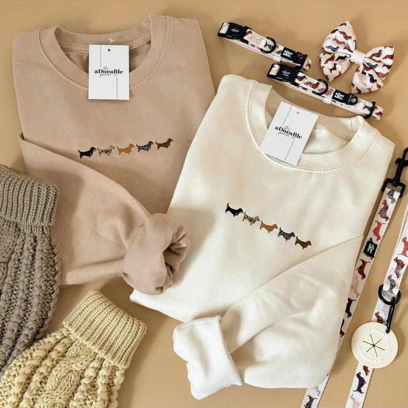 Two folded crewneck sweatshirts from alishabelle, one in beige and one in cream, both featuring a small embroidered line of dachshund dogs on the chest. The sweatsuits are styled in a flat lay with matching accessories including a patterned dog leash, dog collar, dog clip, and dog toy on a warm beige background.