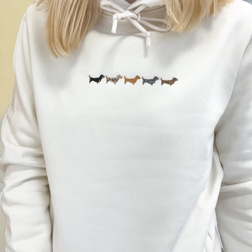 A close-up of a person wearing a white hoodie with a small embroidered row of dachshund dogs across the chest in black, gray, brown, and tan.