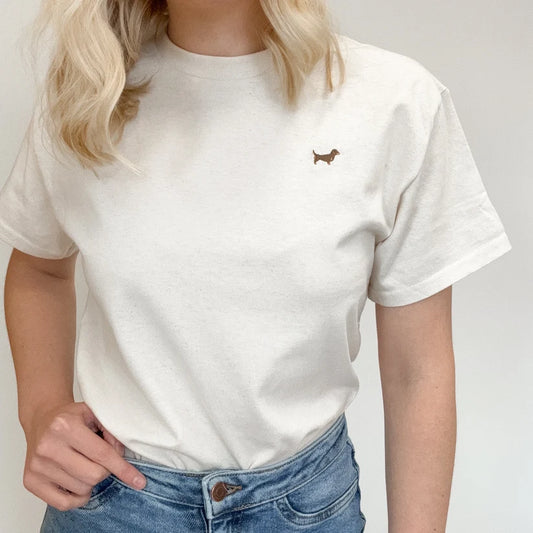 A woman wearing a cream-colored short-sleeve t-shirt with a small brown dachshund embroidered on the left chest, paired with blue jeans.