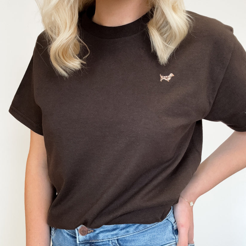 A person wearing a dark brown short-sleeve crewneck t-shirt with a small embroidered dog logo on the left chest, paired with blue jeans.