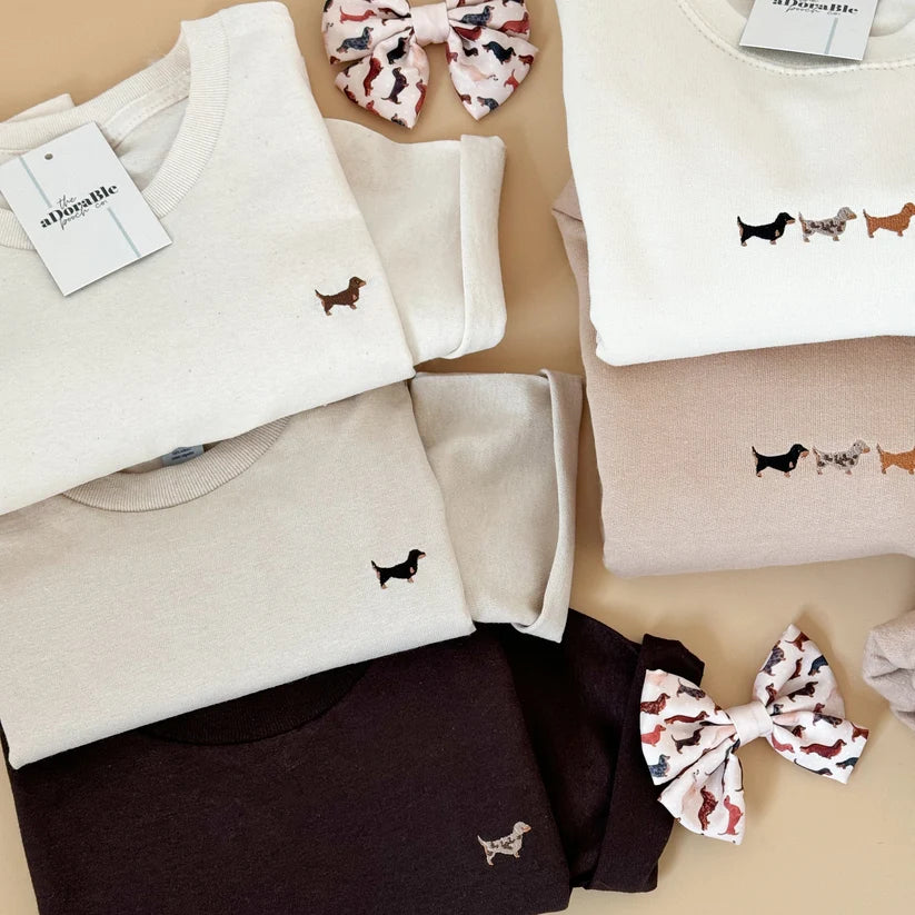 A styled flat-lay of folded crewneck sweatshirts in cream, beige, and dark brown, each featuring a small embroidered dachshund graphic. Two patterned fabric bows are also placed on a warm beige background.