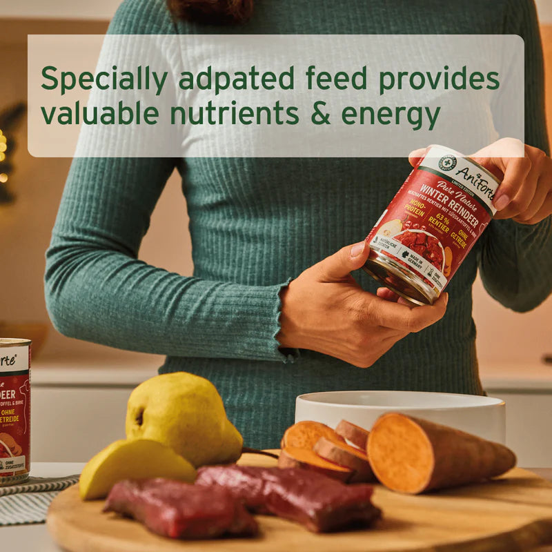 Person holding a can of AniFur pet food with text about specially adapted feed, surrounded by raw meat and vegetables on a table.