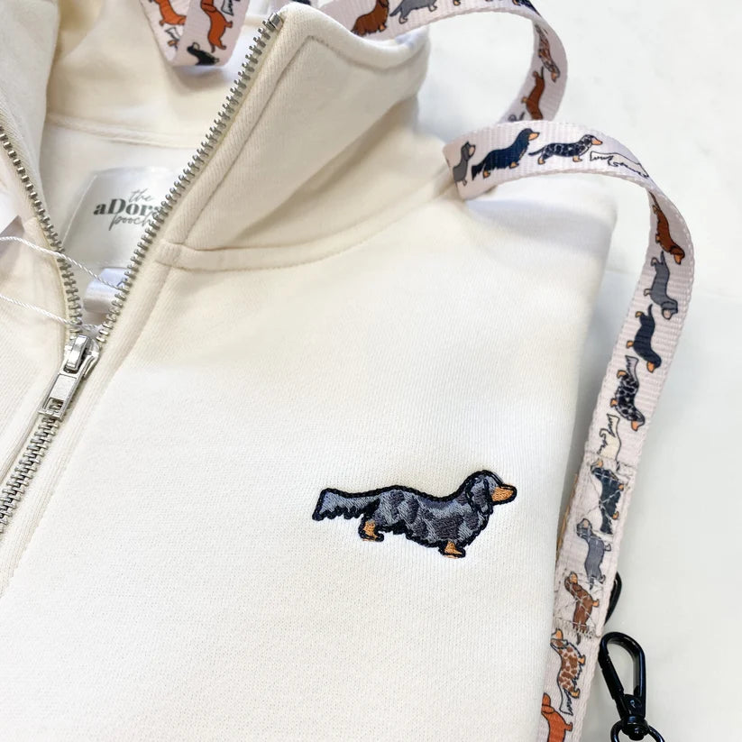 Adorable Pooch cream-colored zip-up hoodie featuring an embroidered black and tan dachshund on the chest, with a matching patterned shoulder strap and a white neck label reading 'aDora'.