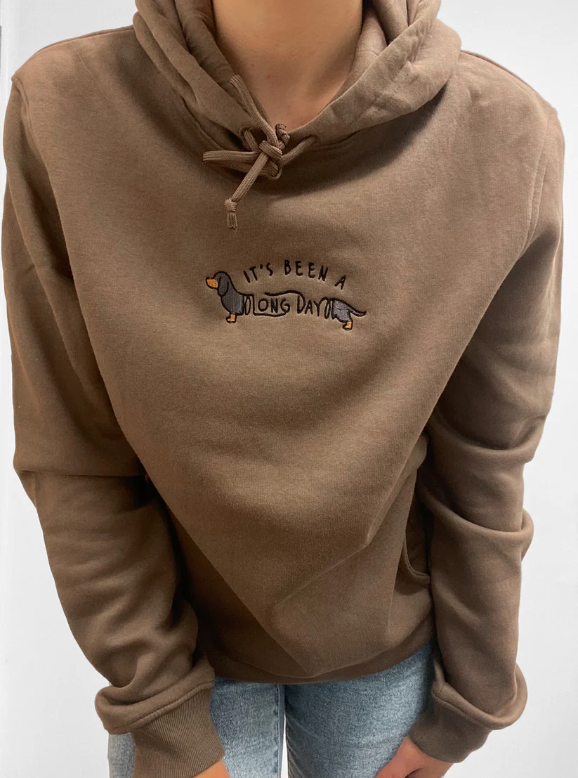 A person wearing a brown hooded sweatshirt with a small embroidered dachshund graphic and the text 'IT'S BEEN A LONG DAY' on the chest, paired with light blue jeans against a plain light background.