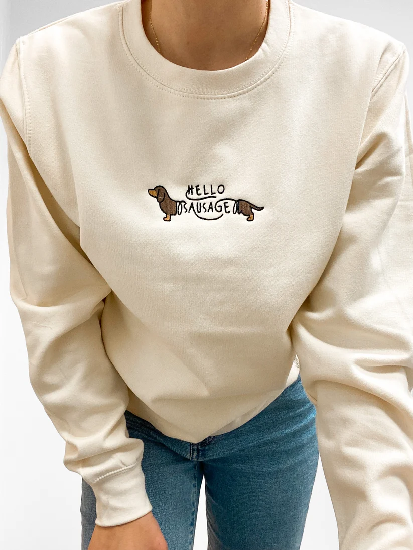 A person wearing a cream-colored crewneck sweatshirt with a small graphic on the chest featuring a dachshund and the text 'HELLO DACHSHUND'. The person is also wearing blue jeans, shown from the neck down against a plain light background.