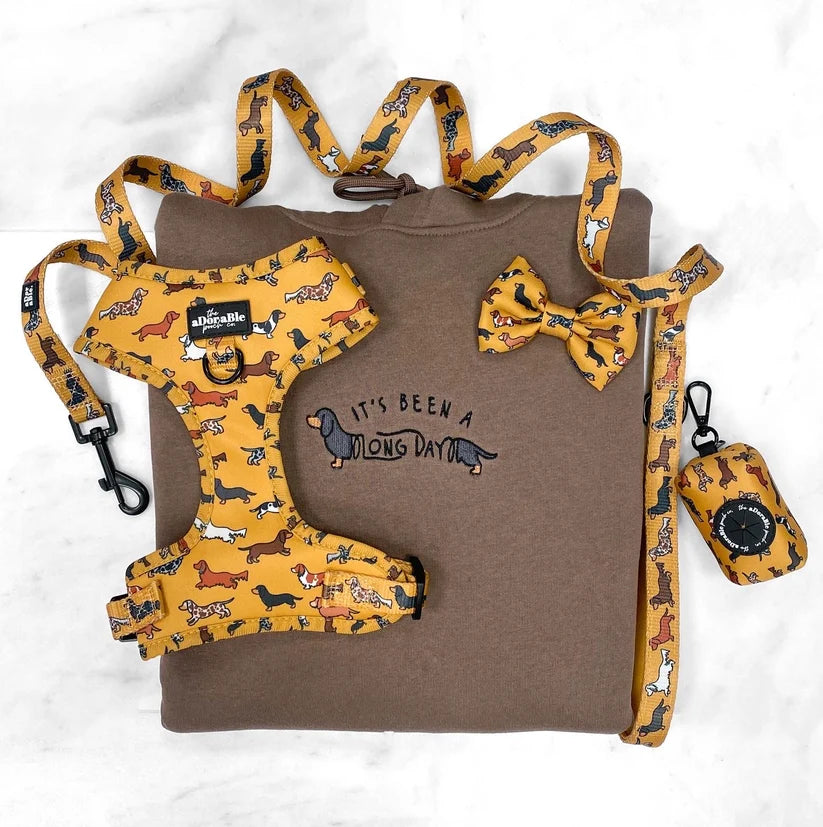 A coordinated pet accessory set laid flat on a white marble-like surface: a brown T-shirt with printed text, a yellow patterned dog harness, matching leash, bow tie, and small bag. The harness and accessories feature a multicolor dog print on a mustard-yellow base.