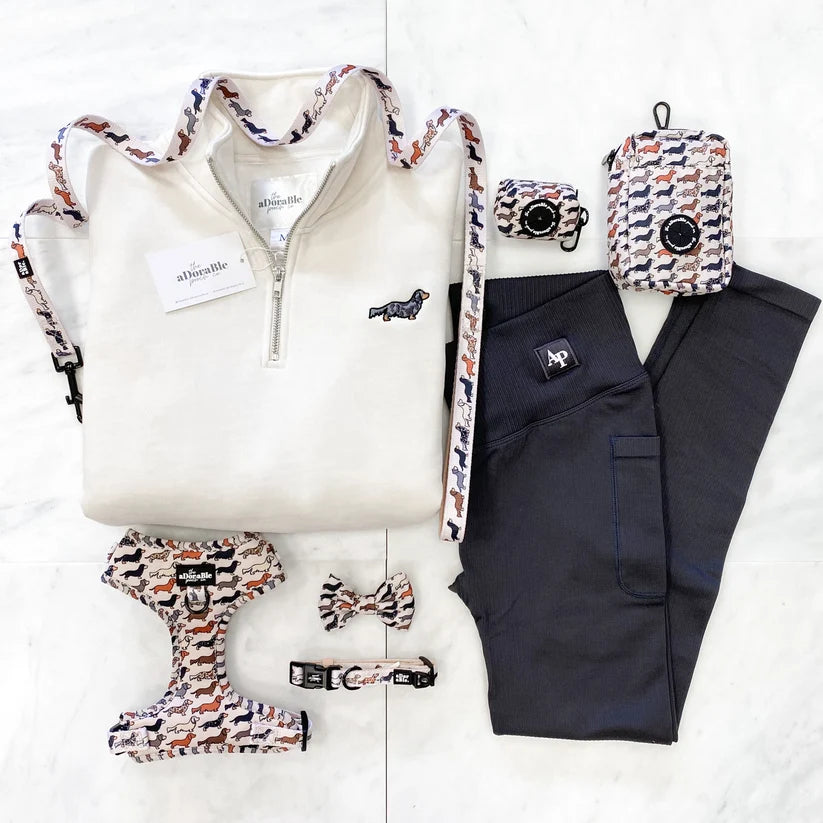 Adorable Pooch coordinated pet apparel set laid flat on a light marble surface, including a cream quarter-zip pullover with patterned trim, dark charcoal pants, a matching patterned harness, small harness bow tie, and matching patterned bag or pouch. The garments and accessories feature a small-scale abstract print in cream, navy, rust orange, and white, with black hardware.
