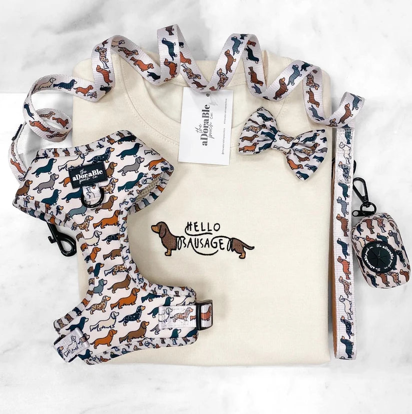 A flat-lay of a dog gear set featuring a cream-colored t-shirt with a dachshund graphic, a matching patterned harness, collar, leash, and small bag, all with a dachshund dog print on a white marble background.