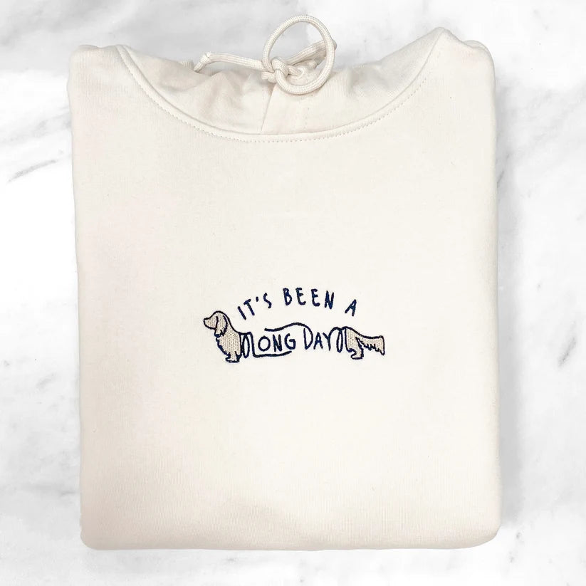 A folded off-white sweatshirt with a drawstring hood, featuring a navy blue graphic of a dachshund and the text 'IT'S BEEN A LONG DAY' on the chest, displayed on a white marble surface.