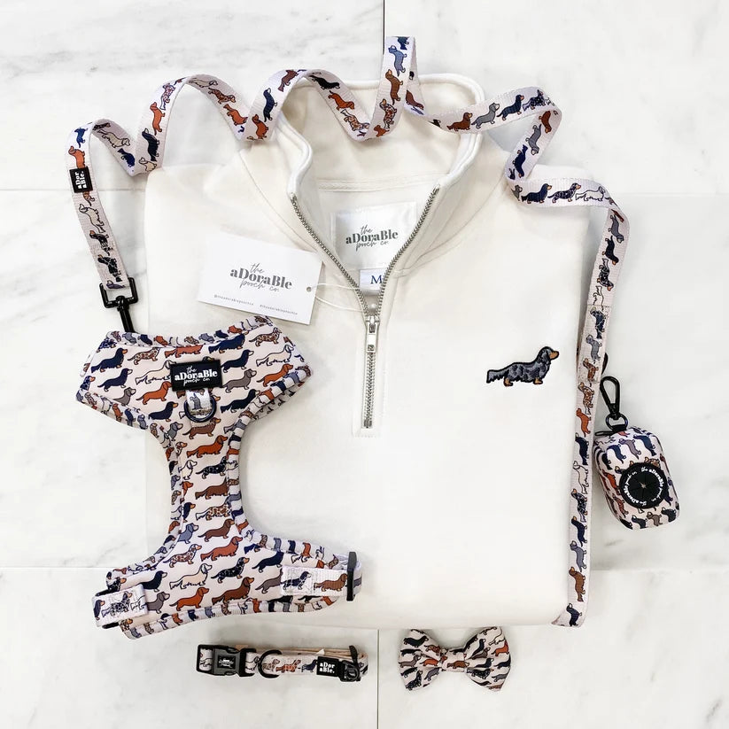 A coordinated dog apparel set laid flat on a white marble surface, including a white quarter-zip pullover sweatshirt, matching harness, leash, collar, bow tie, and small bag, all in a navy, rust orange, and cream pattern. The brand name 'The aDissable Pet' is visible on the neck label, tags, and hardware.