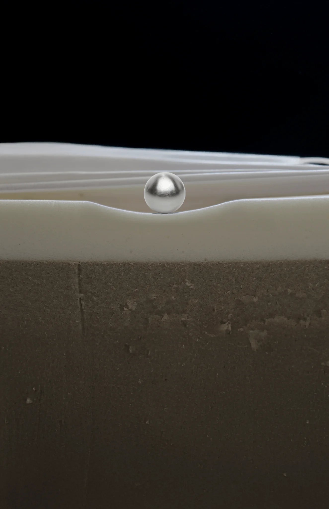 A close-up studio shot of a metallic silver ball resting in a curved valley formed by a thick, off-white layer on top of a textured dark gray-brown material, set against a solid black background.